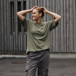 Person wearing a green shirt and gray pants standing outdoors against a wooden wall.