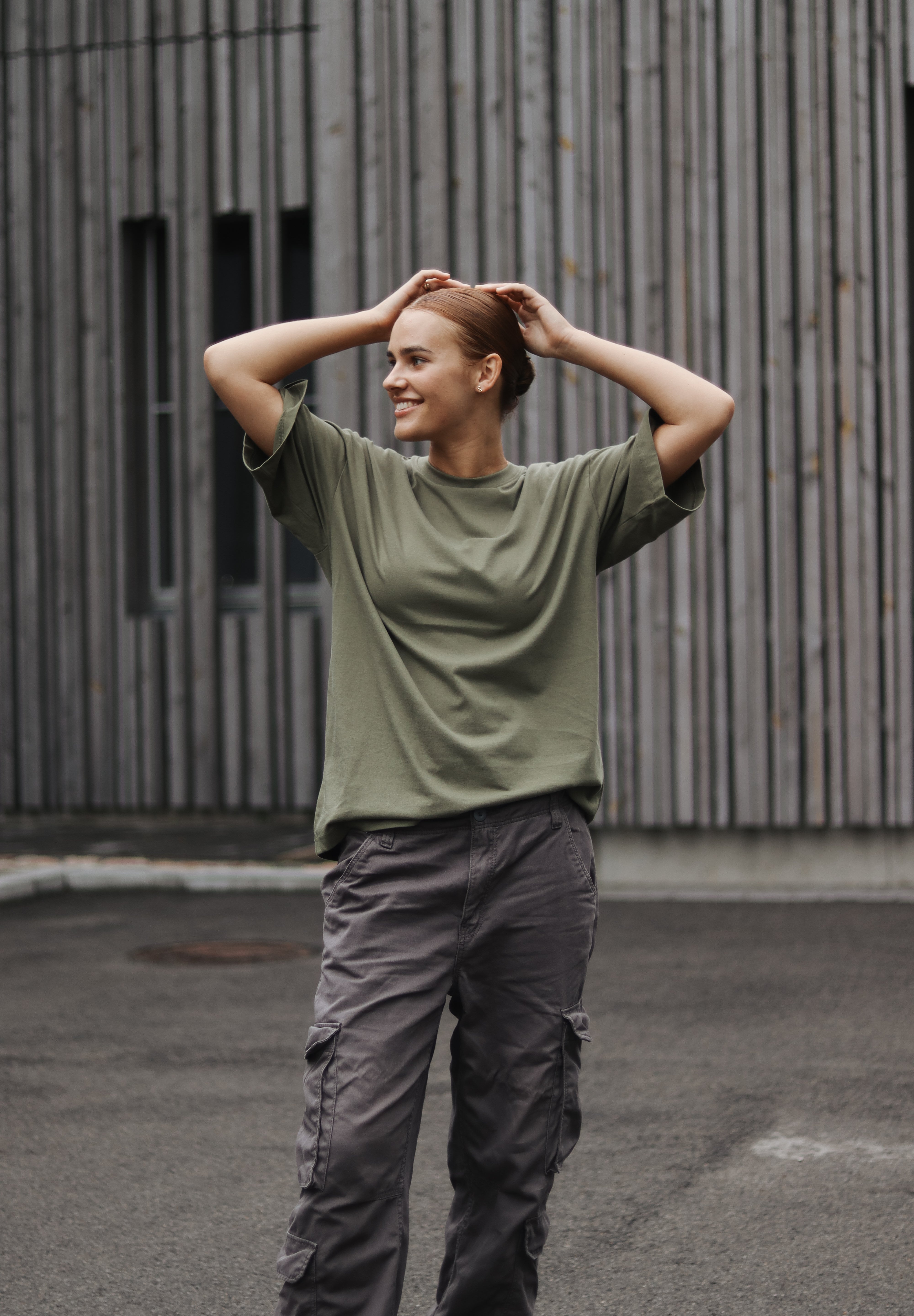 Person wearing a green shirt and gray pants standing outdoors against a wooden wall.