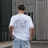 Person wearing a white t-shirt with 'WEAR LOVE' text, standing in front of a modern building.