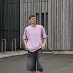 Person wearing a light purple t-shirt and dark shorts standing in front of a modern building.