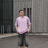 Person wearing a light purple t-shirt and dark shorts standing in front of a modern building.